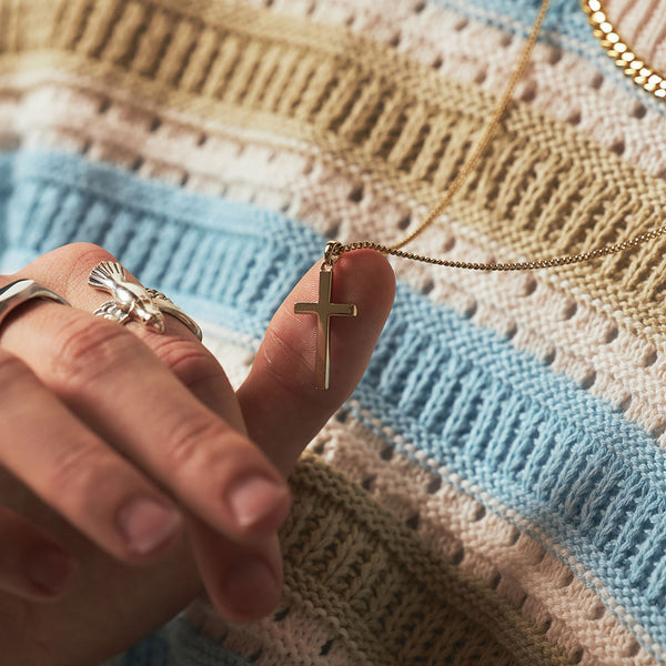 Person wearing silver rings holds the gold cross pendant with their thumb